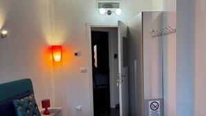 Une chambre d’hôtel comprenant un lit, une table de chevet, une lampe, un luminaire fixé au mur, une porte et un porte-manteau.