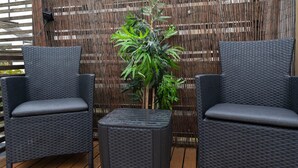 Twee rieten stoelen en een kleine tafel op een houten terras, met een bamboe hek op de achtergrond.
