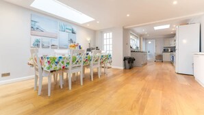 Een ruime eethoek met een lange tafel en stoelen, een keuken op de achtergrond, en een lichte, goed verlichte kamer met houten vloeren.