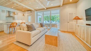 Ein modernes Wohnzimmer mit Sofa, Couchtisch und Essbereich. Es gibt eine Küche mit weißen Schränken und einem großen Fenster.