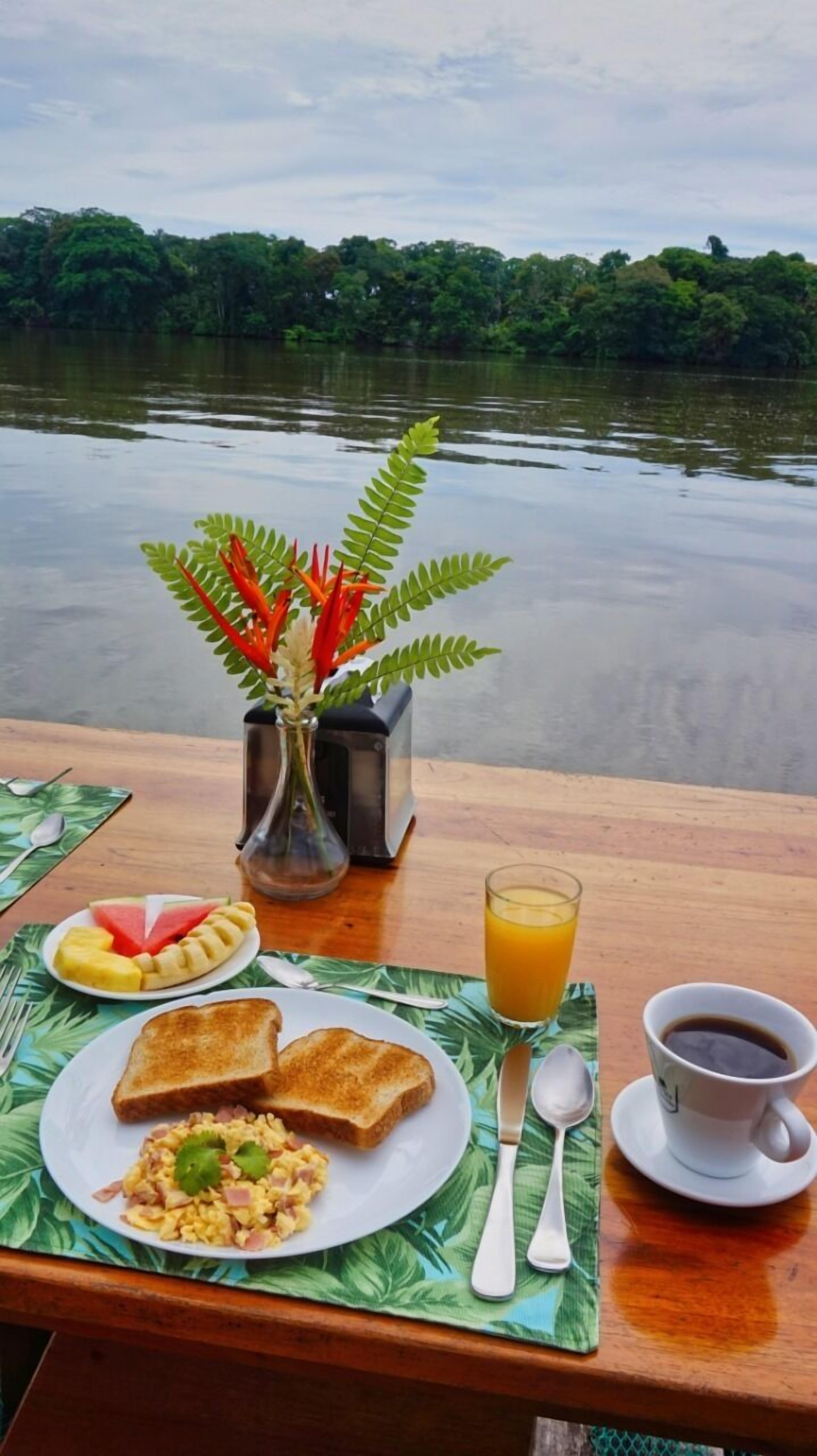 Una mesa de desayuno con tostadas, huevos revueltos, ensalada de frutas, un vaso de jugo de naranja y una taza de café, con vistas a un lago y vegetación.