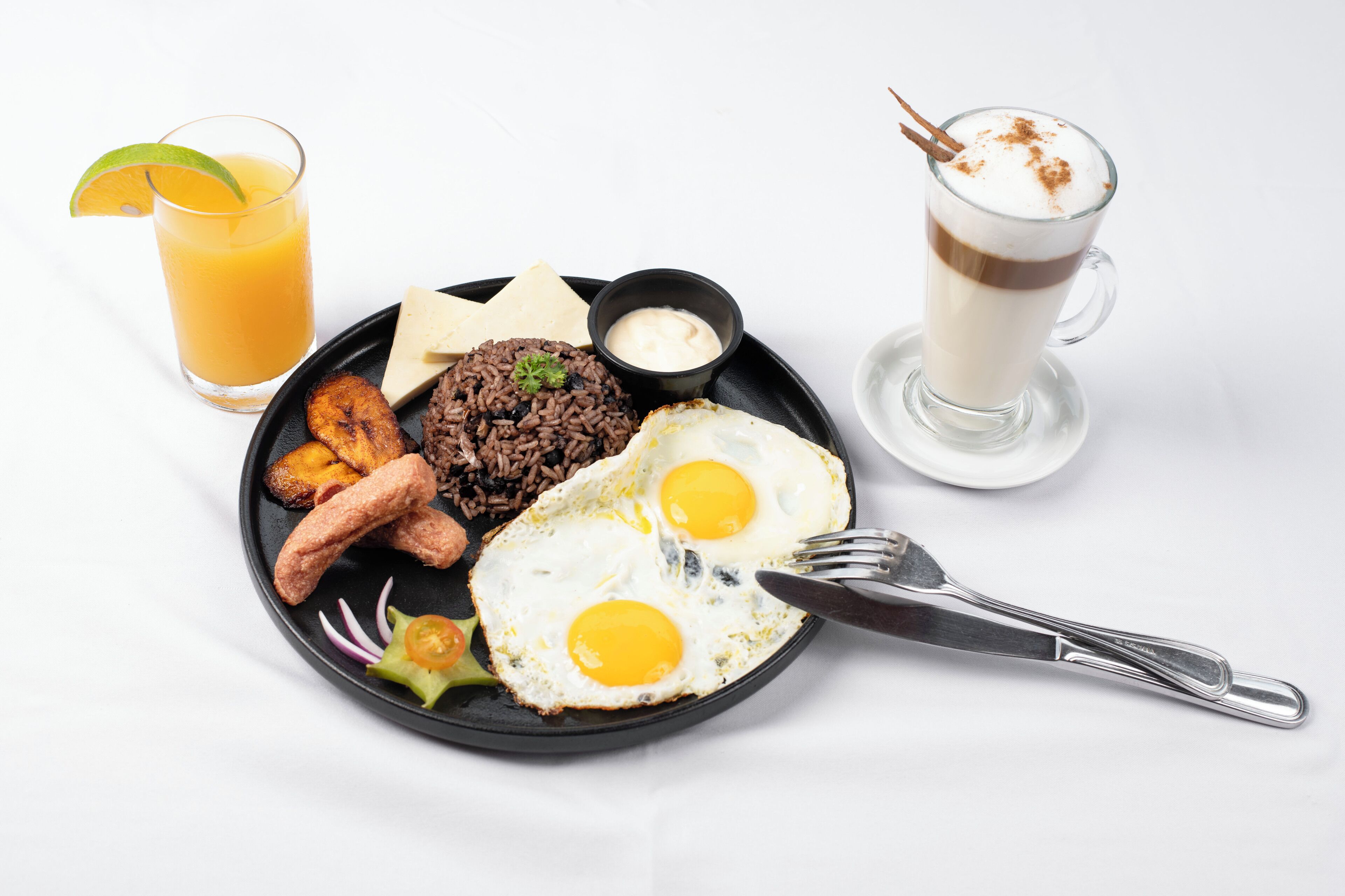 A plate of food including fried eggs, black rice, plantains, and a drink with a slice of lime.