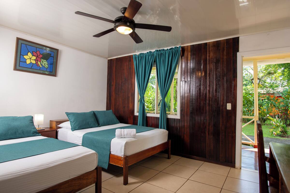 Ein Hotelzimmer mit zwei Betten, einem Deckenventilator, einem Fenster mit Vorhängen und einer Tür zu einem Außenbereich.