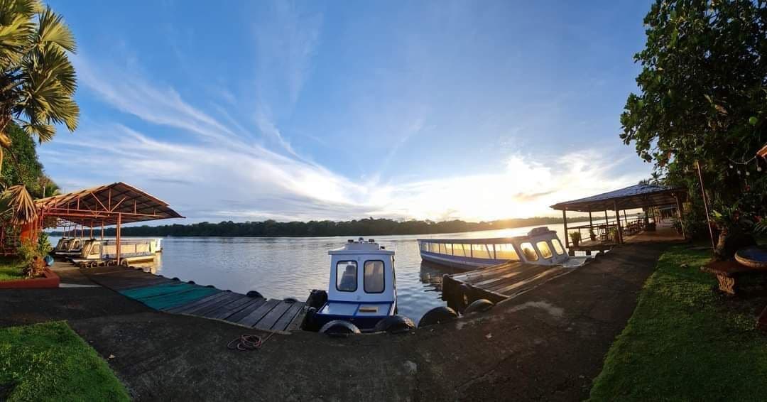 A lakeside dock with a small boat, a covered seating area, and a building with a roof.