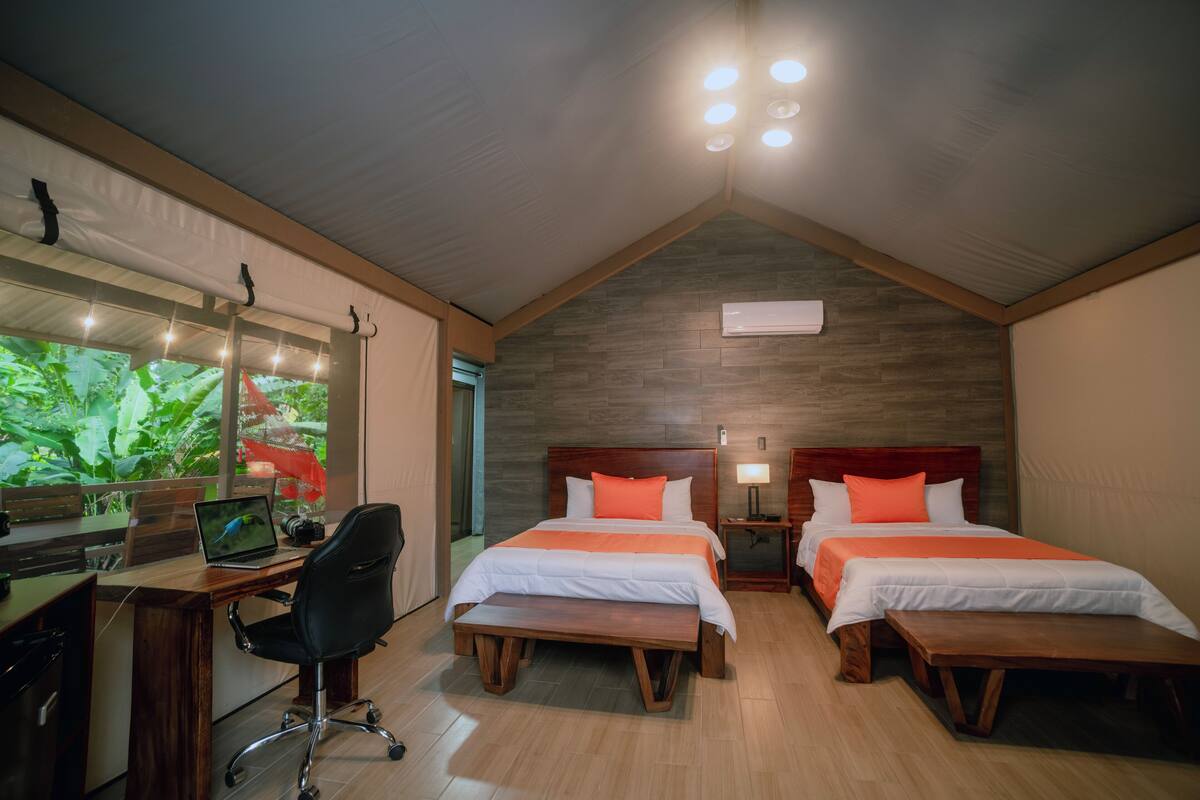 Ein Schlafzimmer mit zwei Betten, einem Schreibtisch mit einem Laptop und einem großen Fenster mit Blick ins Grüne.