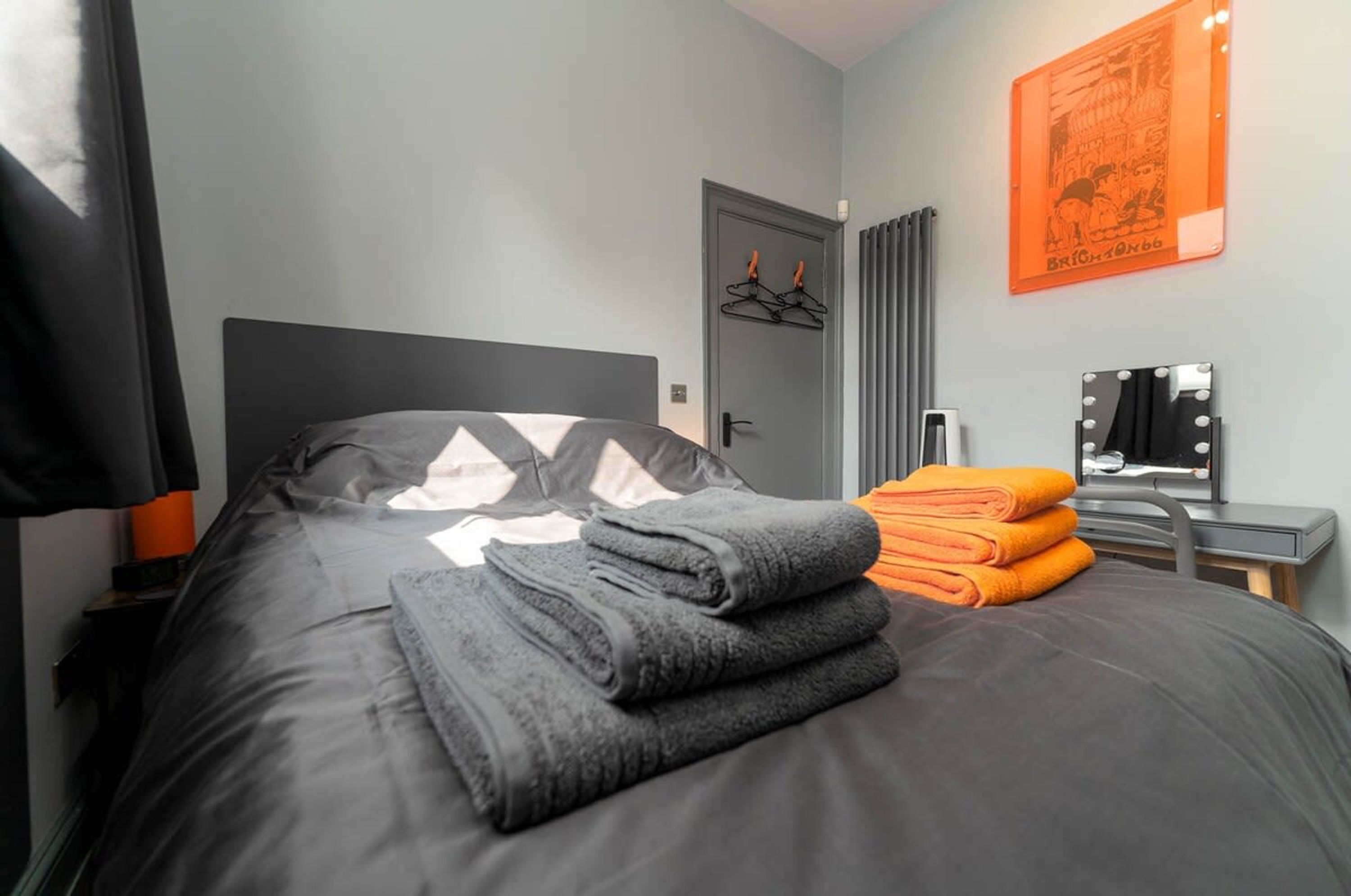 Una cama bien hecha con mantas y toallas dobladas, una puerta con perchero y un espejo con luces.