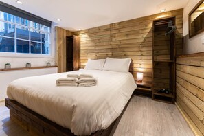 Een slaapkamer met een houten hoofdeinde, een groot bed met witte lakens, een nachtkastje met een lamp en een raam met uitzicht op gebouwen.