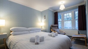 Un lit bien fait, avec du linge de lit et des serviettes blanches, une chaise, une table de chevet et une fenêtre avec des rideaux.