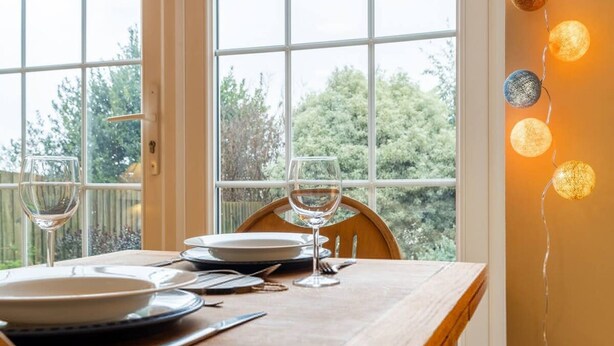 Une table à manger est dressée avec des assiettes, des couverts et des verres à vin, et est placée près d’une fenêtre donnant sur un paysage verdoyant.