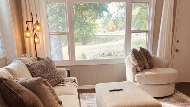 Ein Wohnzimmer mit einem beigefarbenen Sofa, einem Hocker und einem Sessel. Drei Stehlampen und ein großes Fenster mit Blick auf Grünflächen.