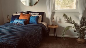 Ein Schlafzimmer mit einem Bett, einem runden Spiegel, einem kleinen Tisch, einer Zimmerpflanze und einem Fenster mit Vorhängen.