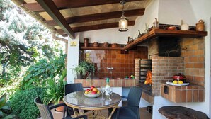 An outdoor patio with a brick oven, wicker chairs, a round table with a bowl of fruit and wine glasses, and a hanging light fixture. - Can Piña - Two Bedroom Villa, Sleeps 4 (Mallorca)