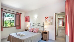 A bedroom with a bed, two bedside tables with lamps, a window with curtains, a bathroom visible through an open door, and a wall with a floral painting. - Can Piña - Two Bedroom Villa, Sleeps 4 (Mallorca)
