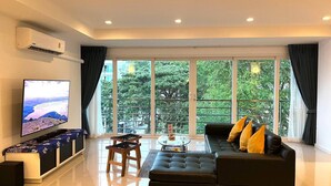 A modern living room with a large TV, a sofa with cushions, a coffee table, and a view of greenery through large windows. - 6-58 - 2 Bdr Spacious Penthouse With Shared Pool (Patong)