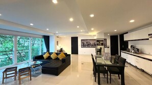 A modern living room with a black sofa, yellow cushions, a glass dining table, and a kitchen area with white cabinets. - 6-58 - 2 Bdr Spacious Penthouse With Shared Pool (Patong)