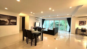 A spacious living area with a dining table, chairs, a sofa, and a TV. There are large windows offering a view of greenery. - 6-58 - 2 Bdr Spacious Penthouse With Shared Pool (Patong)
