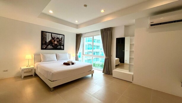 A modern hotel room with a large bed, a bedside table with a lamp, a large window with curtains, and an air conditioning unit. - 6-58 - 2 Bdr Spacious Penthouse With Shared Pool (Patong)