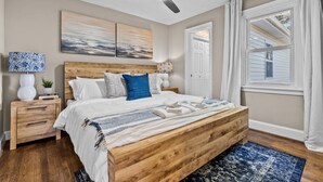 Un dormitorio con una cama de madera, un ventilador de techo, dos obras de arte en la pared, una mesita de noche con lámpara y una ventana con cortinas.