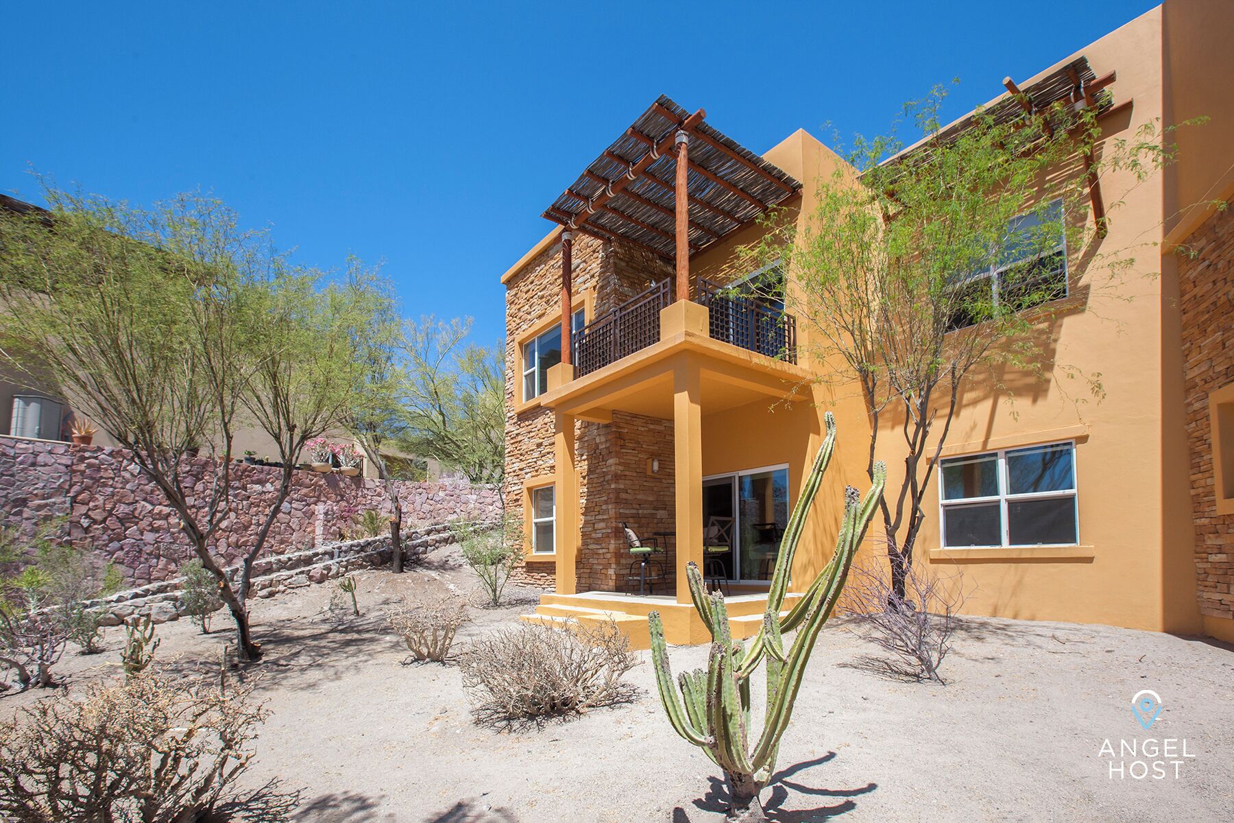 Un edificio de dos pisos con fachada de piedra, un balcón con pérgola de madera y un jardín desértico con cactus y árboles pequeños.