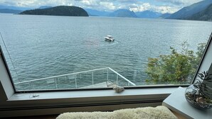 A room with a large window overlooking a lake, a small bench, and a potted plant on a side table. - Pacific Gem 💎 Ocean Front  (West Vancouver)