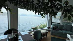 A modern living room with a large window overlooking a body of water, a dining area with a table and chairs, and a cozy seating area with a sofa. - Pacific Gem 💎 Ocean Front  (West Vancouver)