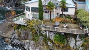 A coastal house with a glass conservatory, outdoor seating, and a rocky cliff backdrop. - Pacific Gem 💎 Ocean Front  (West Vancouver)