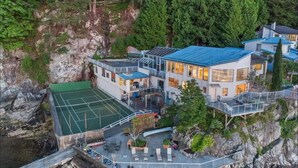 A modern multi-story building with a tennis court, outdoor seating, and a view of a rocky cliff and forest. - Pacific Gem 💎 Ocean Front  (West Vancouver)