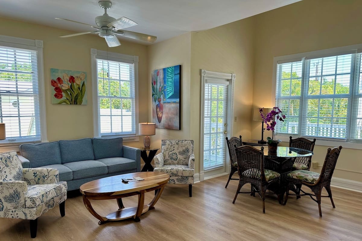 A living room with a sofa, coffee table, and dining table with chairs. There are windows with blinds, a ceiling fan, and artwork on the walls.