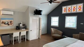 A bedroom with a bed, a desk with chairs, a ceiling fan, and a TV.