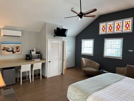 A bedroom with a bed, a desk with chairs, a ceiling fan, and a TV.