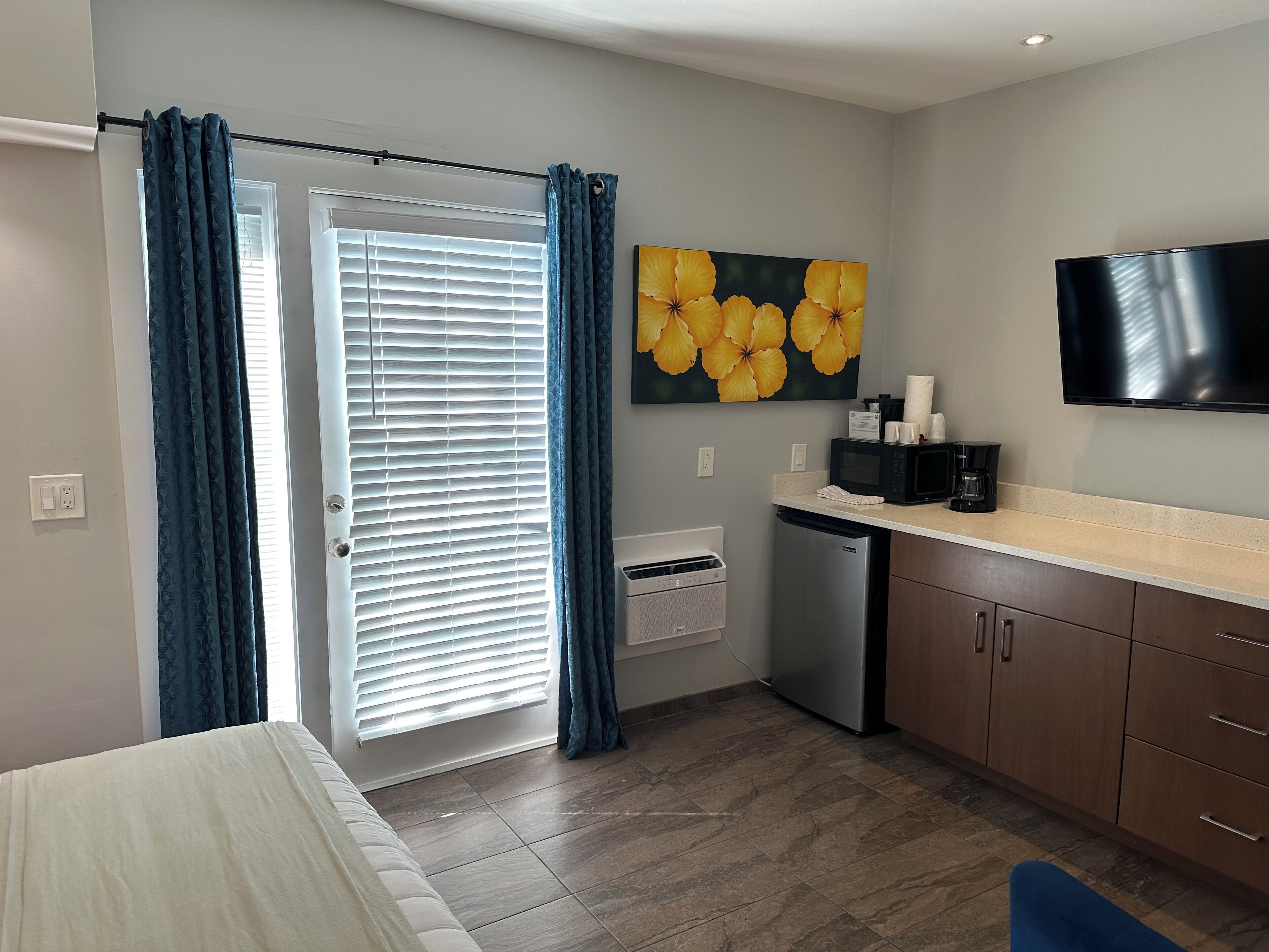 Ein modernes Hotelzimmer mit Bett, einem Badezimmer mit Dusche, einer Küchenzeile inklusive Mikrowelle und Kaffeemaschine sowie einem großen Fenster mit Vorhängen.