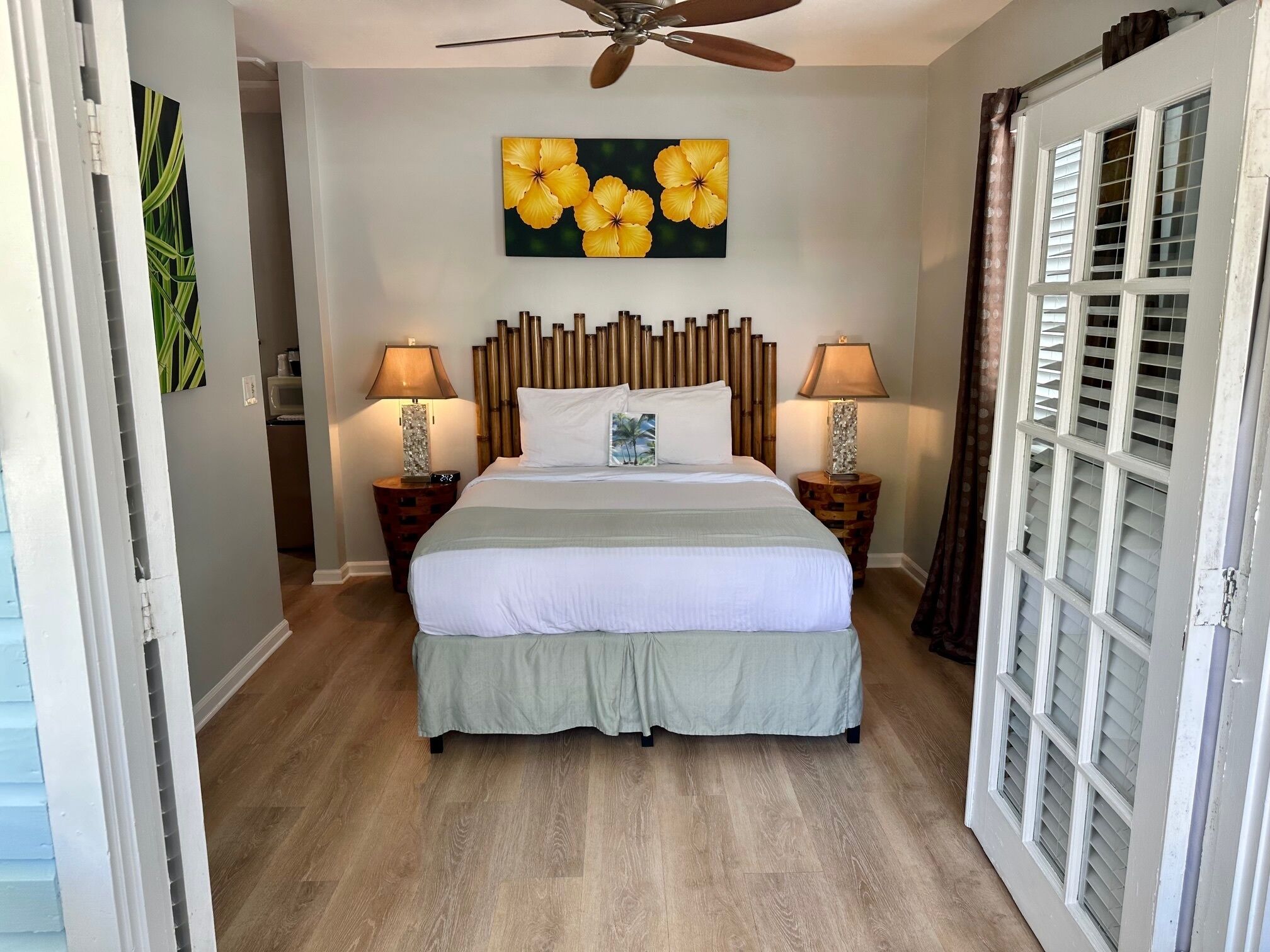 Quarto de hotel com cama, duas mesas de cabeceira com abajures, um ventilador de teto, uma parede com pintura floral e uma porta de vidro deslizante.