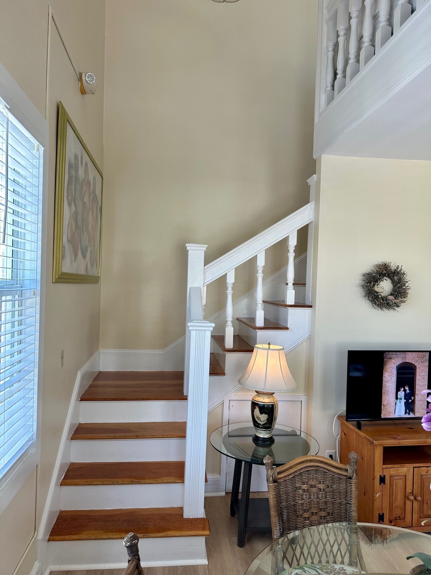 A staircase with white balusters, a glass table with a rooster lamp, and a television displaying a wedding scene.