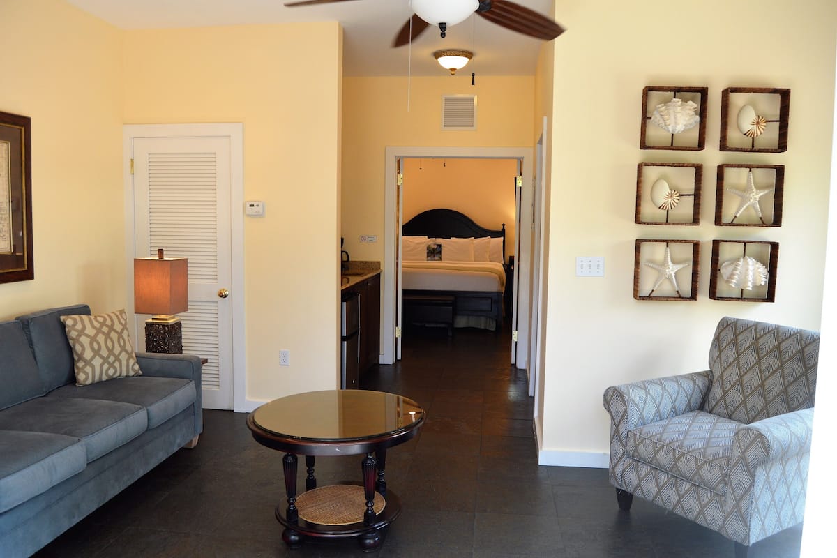 A living room with a sofa, armchair, and a round table. There is a door leading to a bedroom with a bed and a ceiling fan.