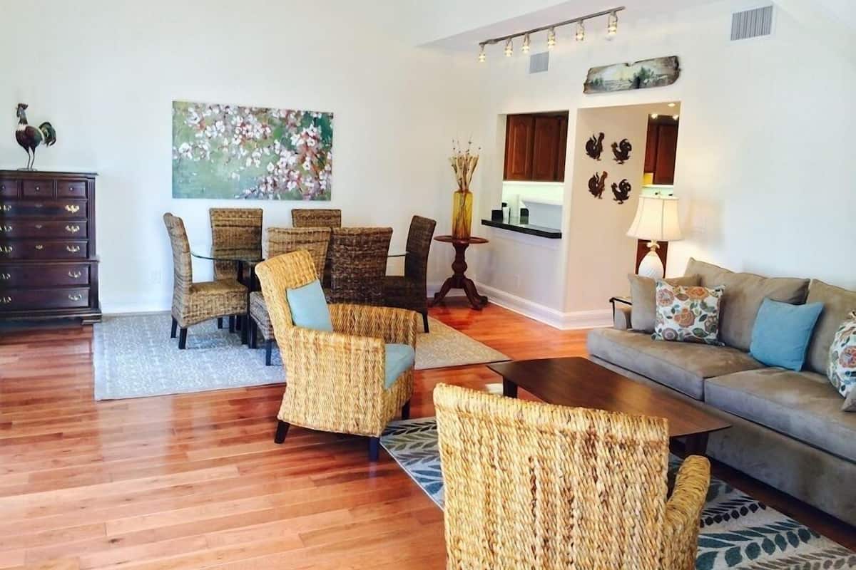 A spacious living room with a high ceiling, a dining area with wicker furniture, and a kitchen visible through an open doorway.