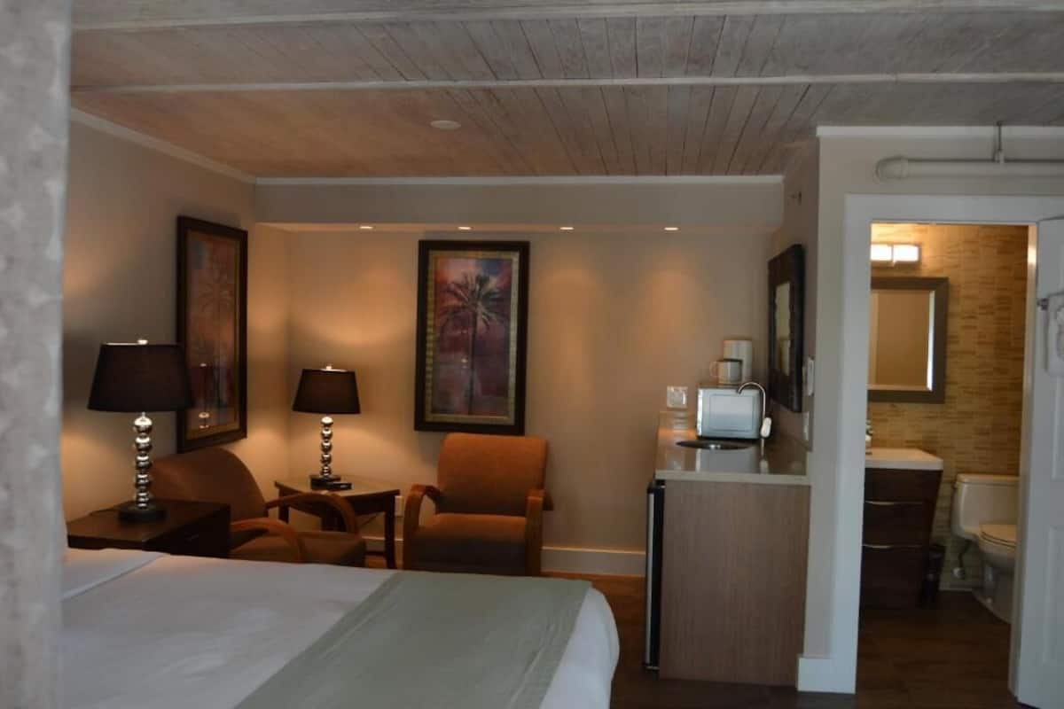 A hotel room with a bed, two chairs, a desk with a lamp, a painting, a television, and a bathroom visible through an open door.