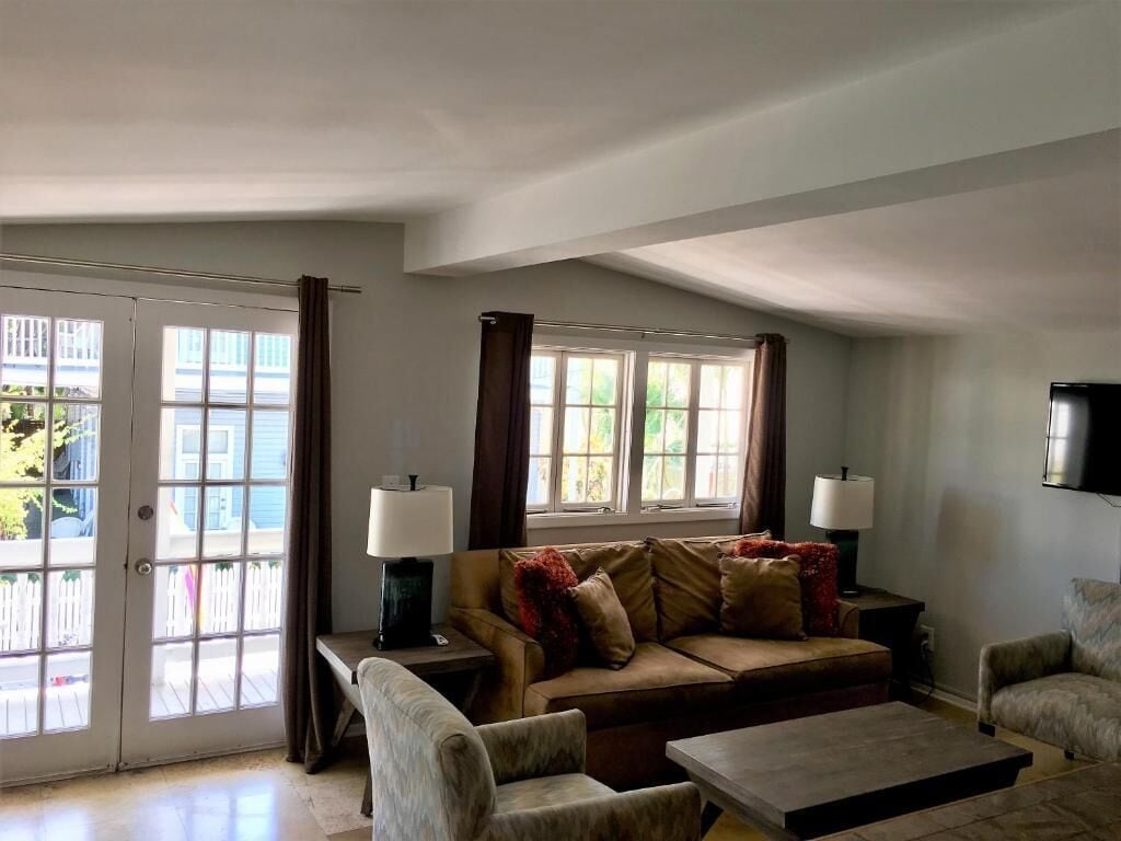 A living room with a sofa, armchairs, a coffee table, and two lamps. There are two windows with curtains, a door, and a flat-screen TV mounted on the wall.