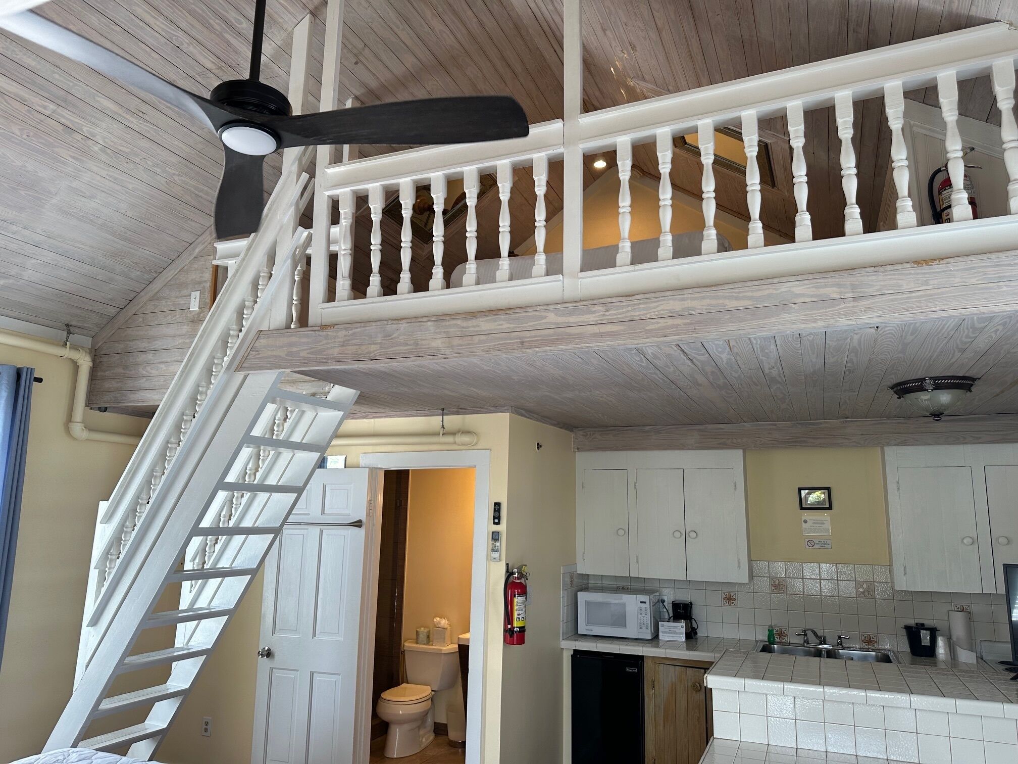 Eine geräumige Küche mit weißen Schränken, einer Mikrowelle und einem Spülbecken. Eine weiße Treppe führt zu einer oberen Ebene mit Holzboden und Deckenventilator.