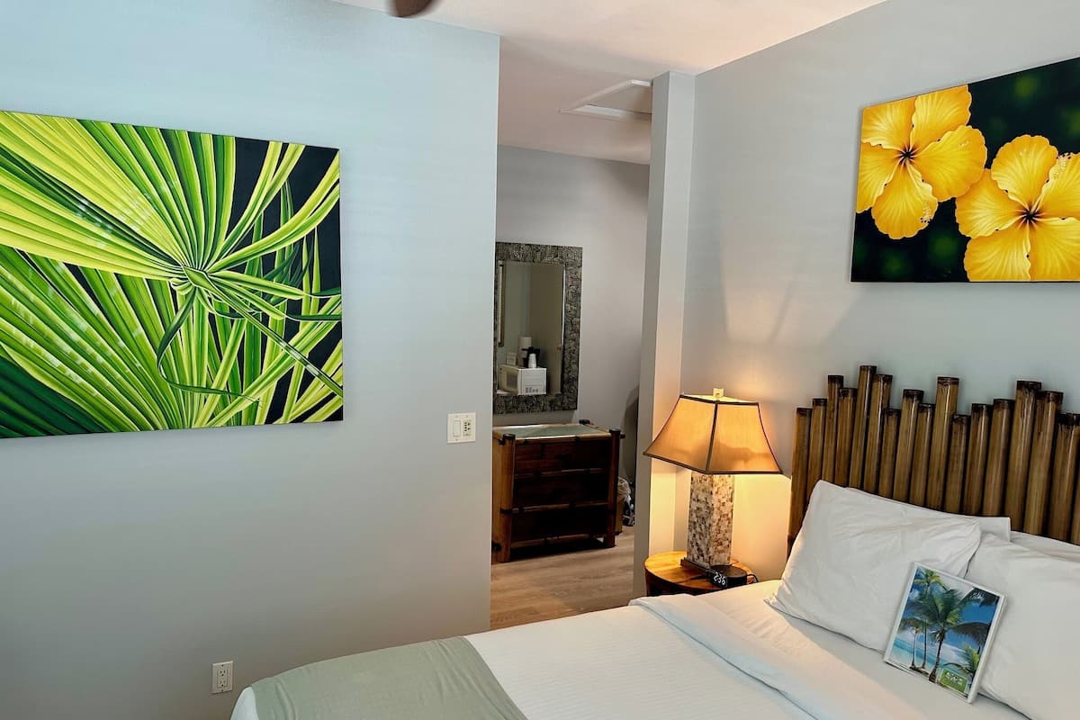 A hotel room with a bed, a nightstand, a lamp, a mirror, and a painting of yellow flowers on the wall.