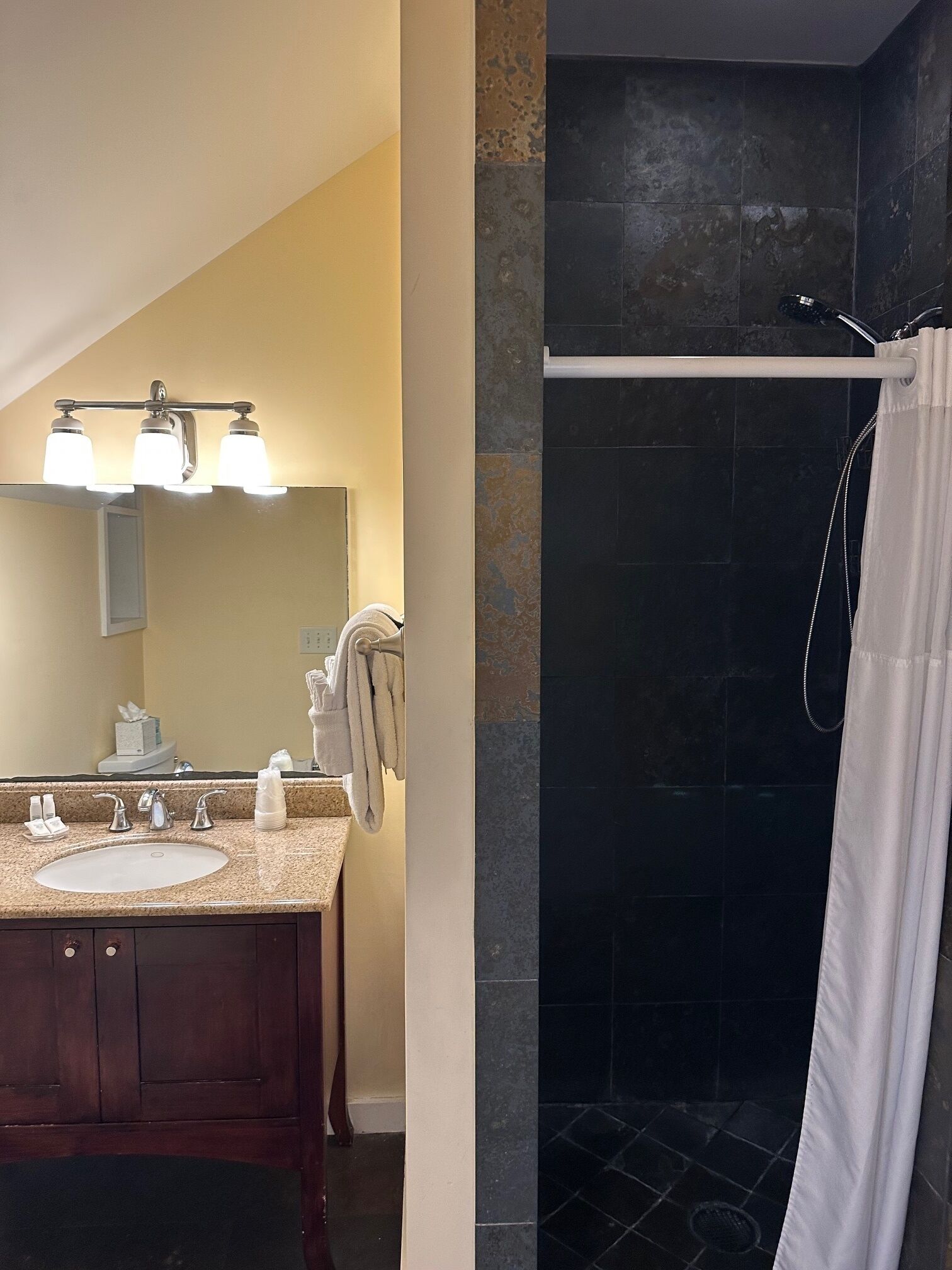 A bathroom with a vanity, sink, and mirror. There is a shower with a curtain and a towel hanging on the wall.