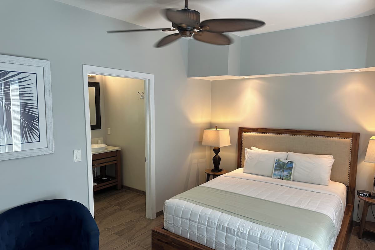 A bedroom with a bed, bedside lamps, a ceiling fan, and a bathroom visible through an open door.
