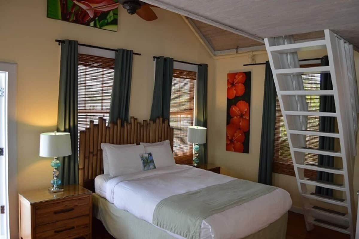 A bedroom with a bed, two bedside tables with lamps, a wooden headboard, a ladder, and a large window with blinds.