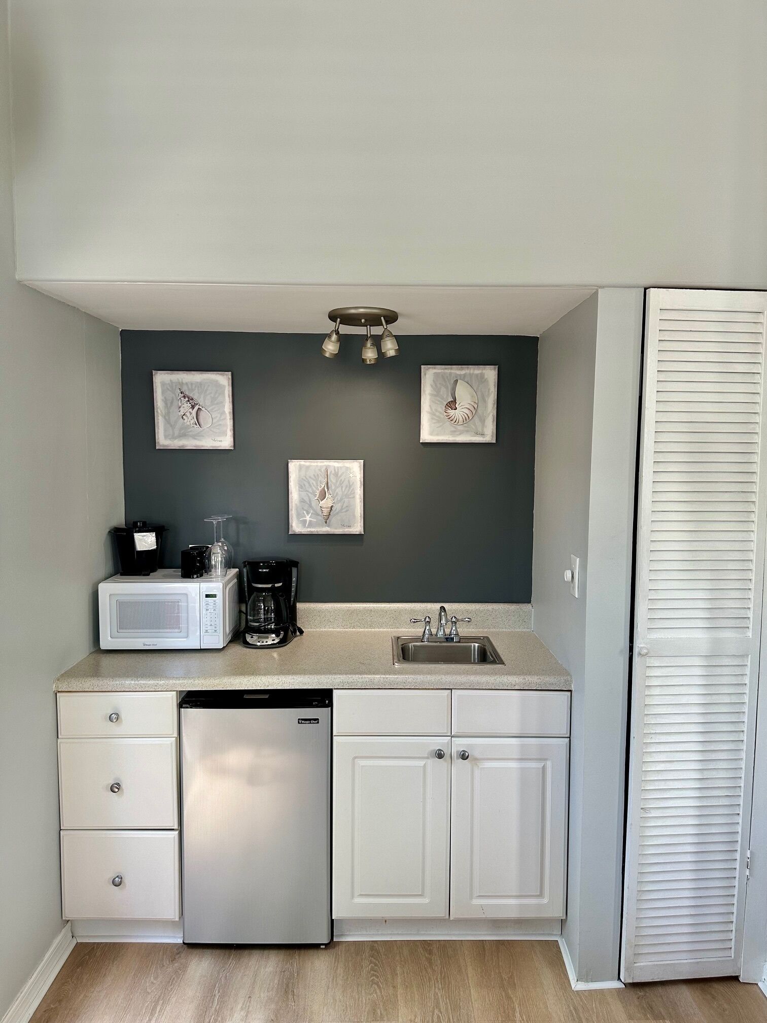 A compact kitchen with a microwave, coffee maker, and sink. There are three framed pictures on the wall, a white cabinet, and a white door with louvered panels.