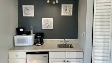 A compact kitchen with a microwave, coffee maker, and sink. There are three framed pictures on the wall, a white cabinet, and a white door with louvered panels.