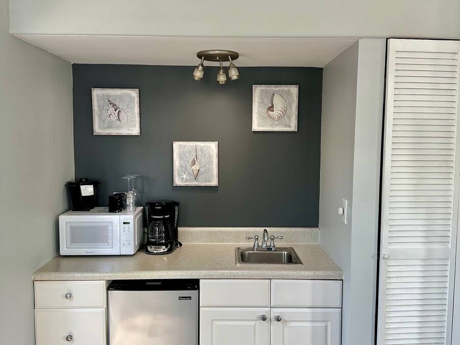 A compact kitchen with a microwave, coffee maker, and sink. There are three framed pictures on the wall, a white cabinet, and a white door with louvered panels.