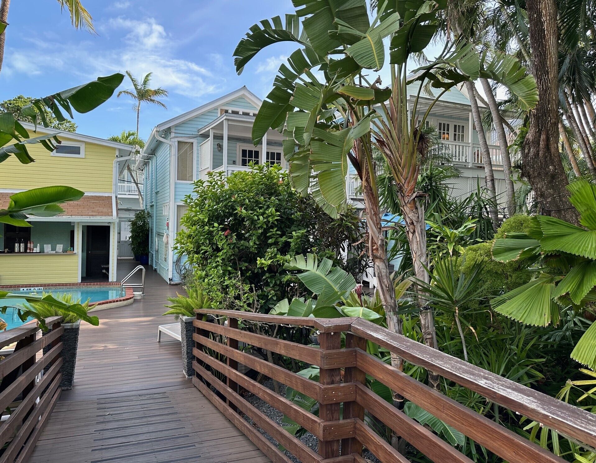 A wooden deck leading to a pool area with tropical plants and a building with a light blue facade.