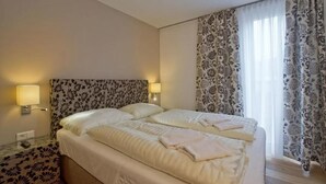 Habitación de hotel con una cama grande, dos lámparas de noche, cortinas con estampado floral y una ventana con cortinas blancas traslúcidas.
