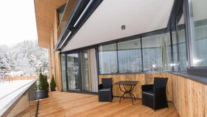 Una terraza de madera con dos sillas y una mesa, ventanas amplias y vista a árboles nevados.