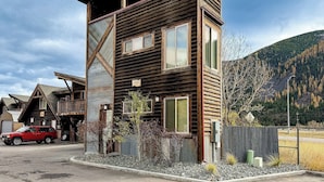 Une cabane en bois à deux étages, avec un toit en pente, une voiture rouge garée devant, et un paysage montagneux en arrière-plan.