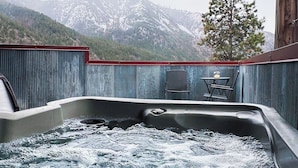 Un bain à remous avec vue sur des montagnes enneigées, une terrasse en bois et une table avec des chaises.